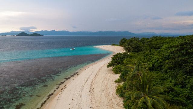 Aerial view of a pristine white sand beach with a small boat anchored in turquoise waters, bordered by lush tropical vegetation and palm trees in Busuanga, Mimaropa, Philippines.