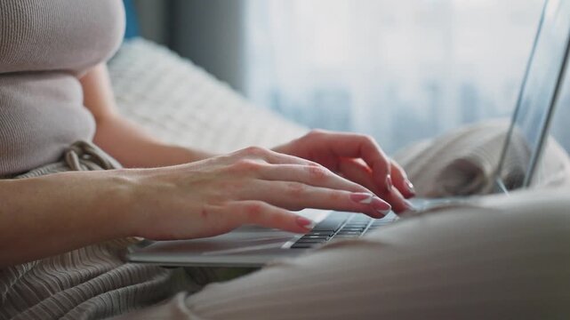 Soft light fills cozy room with gentle blur. Woman's hands type on laptop, calm and focused. Comfortable seating suggests relaxation or remote work. Laptop sits open