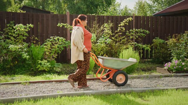 Woman pushes wheelbarrow across garden path. Gravel borders green grass and garden beds. She wears patterned pants, orange top, and light jacket. Her hair is tied back, she walks with purpose