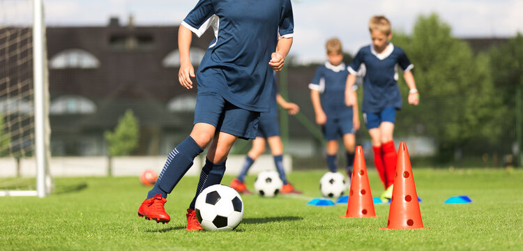 Soccer Outdoor Slalom Cone Drill. Junior Soccer Player in Dark Blue Kit Dribbling Ball Through Orange Cones During Outdoor Training Drills on Grass Pitch