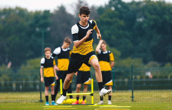 Youth Soccer Player in Yellow and Black Kit Practicing Agility Ladder Jump Drill on Outdoor Pitch During Training Session