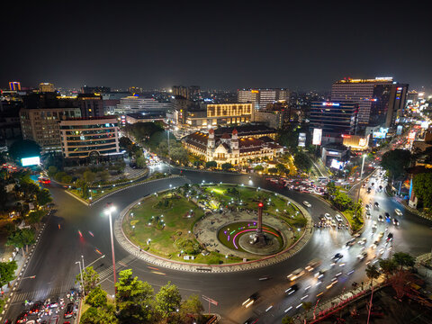 Aerial view of Simpang Lima Semarang roundabout at night with illuminated city buildings, busy traffic light trails, and lush green park in Kecamatan Semarang Tengah, Jawa Tengah, Indonesia.