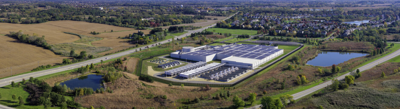 Aerial view of a large data center facility with rooftop solar panels and cooling units, surrounded by autumn fields, roads, and small ponds in Volo, Illinois, United States.