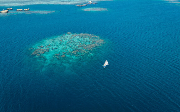 Aerial view of a traditional boat sailing near vibrant coral reefs and floating fish cages in the clear blue waters of Karimunjawa, Central Java, Indonesia.