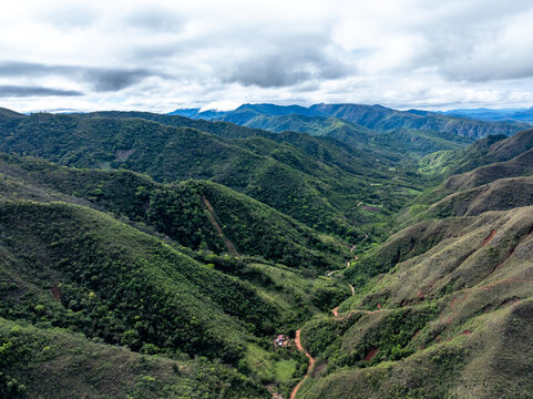 Aerial view of Codo de los Andes featuring lush green mountain slopes, a winding dirt road through the valley, and a cloudy sky in Samaipata, Santa Cruz Department, Bolivia.