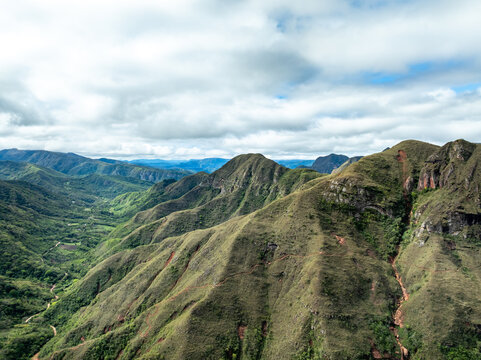 Aerial view of the Codo de los Andes mountain range featuring rugged green slopes, deep valleys, and winding trails under a cloudy sky in Samaipata, Santa Cruz Department, Bolivia.
