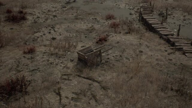 An empty wooden wheelbarrow sits on parched earth by a broken bridge, surrounded by dry grass and mud puddles in a desolate, barren landscape.