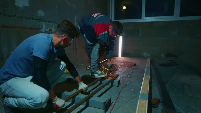 Two workers cut metal with a grinder. Sparks fly as they work inside a dimly lit building. They focus on the task, wearing safety gear. Ideal for construction or industrial projects