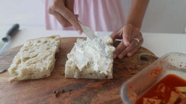 Hands spreading whipped white cheese over rustic bread slices on wooden cutting board in bright kitchen with pale pink apron visible