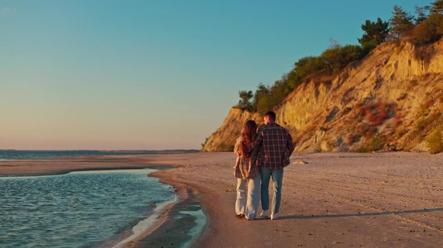 A couple walks along a quiet shoreline beneath sunlit coastal cliffs at golden hour