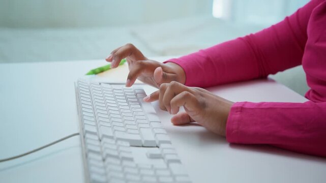 Pink-sleeved hands type on a white keyboard. Fingers move steadily over keys with focused intent. White mouse rests beside the keyboard. Bright, clean workspace under soft daylight