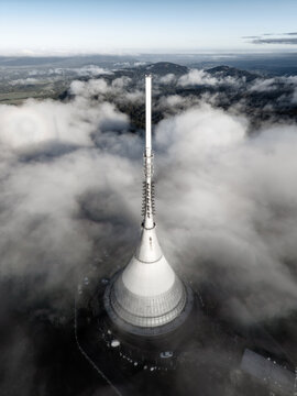 Aerial view of the iconic Jested Tower rising above a thick layer of clouds, showcasing its unique hyperbolic structure and antenna spire in Liberec, Liberecky kraj, Czechia.