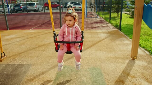 Tiny girl in pink, swinging gently on playground swing. She grips the handle, eyes soft, looking ahead. Sunlight glints off the playground surface, casting warm shadows. Background shows parked cars