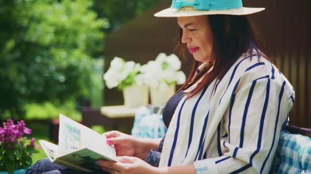 Woman relaxes in garden, reading book. She wears wide-brimmed hat and striped top. Her expression shows calm, contented focus. Lush greenery surrounds her, with soft flowers nearby