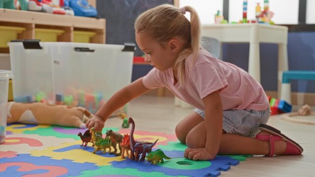 Blonde girl kneels and places toy dinosaurs in a row on a colorful foam mat inside a building playroom, hand reaching to arrange figures; imaginative joy.