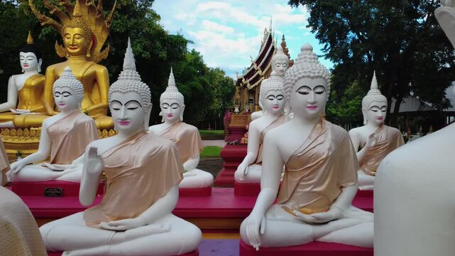 Close-up Row of white and gold Buddha statues seated beside an old brick pagoda in a traditional Thai temple.	