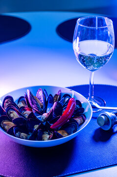 Vertical view of a mussels bowl and a glass of white wine bathed in cool blue light on a white table. Moody evening seafood for wine bar stories and night dinner concept.