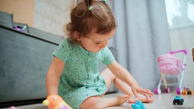 Little girl plays with toy cars on the floor. She wears a green dress with tiny patterns. Her curly hair is held with a clip. She reaches toward a blue vehicle with curiosity