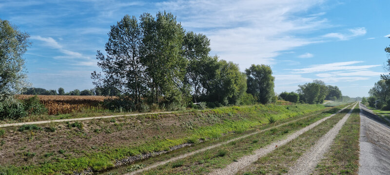 A sunny rural landscape showing a gravel double track road on a water levee next to Varazdin lake with green trees.