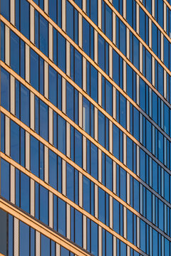 Golden diagonal metal lines cross a reflective blue glass building facade creating an abstract geometric pattern with warm sunlight and regular vertical divisions