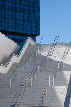 Broad outdoor concrete staircase with repeated metal handrails leading toward blue glass towers under a clear sky in a contemporary urban public space