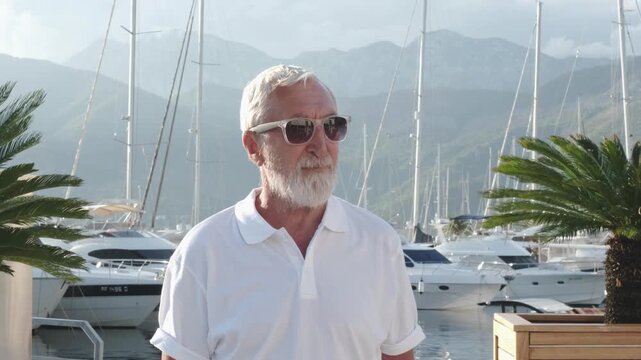 Close up of senior man in sunglasses at sailing marina, grey bearded man in white polo looking at boats with mountains in Tivat, Montenegro. Yacht charter concept, man turns and smiles