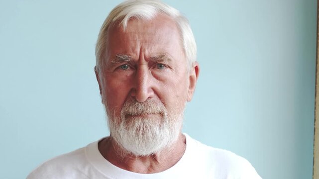 Stern senior man looking at camera, grey bearded elderly man in white t-shirt with serious frowning face on light blue background. Strictness and discipline concept, studio portrait