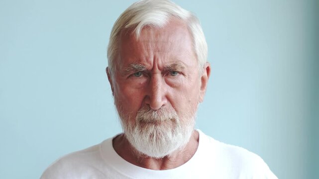 Grumpy old man frowning, grey bearded senior in white t-shirt looking at camera with distrust on light blue background. Bad mood and complaint concept, studio portrait of elderly man