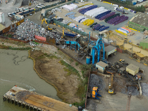 Aerial view of a recycling facility with piles of scrap metal, heavy machinery, and rows of colorful containers near a riverbank in the United Kingdom.