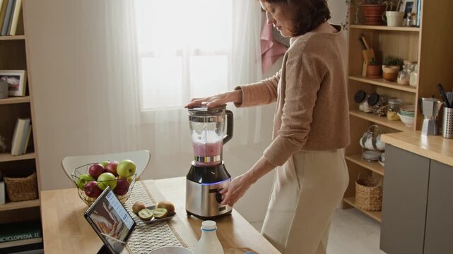 Caucasian middle aged woman blending fruit smoothie in electric blender while following online recipe on tablet. Ideal for healthy nutrition campaigns, cooking apps, appliance advertising