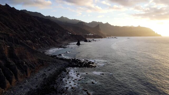 A drone flies low along the sunlit cliffs of the Anaga coast near Benijo with the ocean glowing under a low sun. Dramatic backlit island scenery for travel productions.