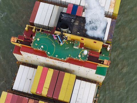 Aerial view of a large cargo ship deck loaded with colorful shipping containers and a helipad, with smoke rising from the funnel in the United Kingdom.