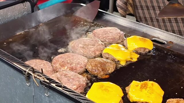 juicy beef burger patties with melting yellow cheese grilling on hot flat iron griddle pan at street food stall