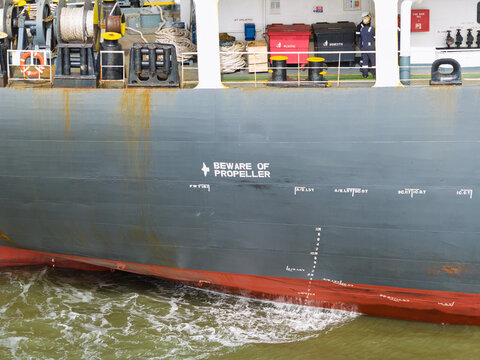 Aerial view of the hull of a large ship featuring a warning sign, draft markings, and deck equipment in the United Kingdom.