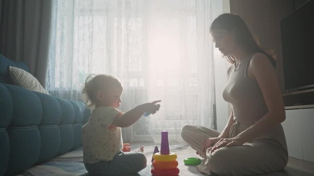 Soft sunlight bathes a cozy living room. Mother and child sit close, engaged in quiet play. Child focuses on stacking colorful blocks with care. Mother watches, smiling gently, offering quiet support