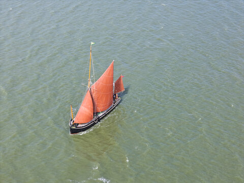 Aerial view of a traditional Thames sailing barge with distinctive red sails navigating through open water in the United Kingdom.