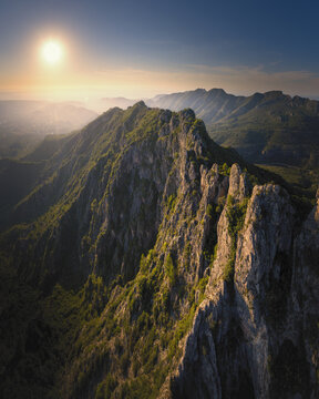 Aerial view of the rugged mountain range at sunset, with rocky cliffs and green vegetation illuminated by golden light in La Barraca d'Aigues Vives, Valencian Community, Spain.