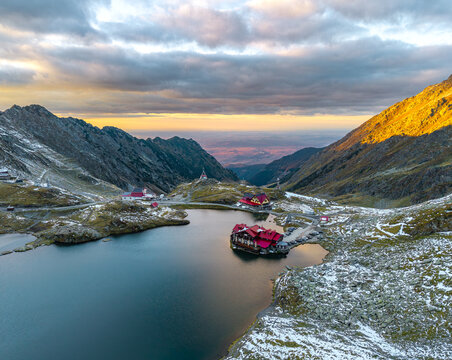 Aerial view of Balea Lake surrounded by rugged mountains, rocky slopes with patches of snow, and red-roofed buildings at sunset in Cartisoara, Sibiu, Romania.