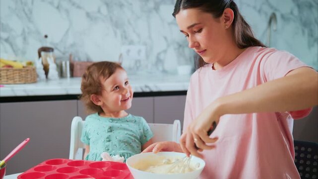 Mom smiles while mixing batter with child. Child watches with joy, seated in high chair. Kitchen features marble backsplash and bright lighting. Perfect for family, cooking, or parenting content