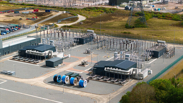 Aerial view of the National Grid Bramford 400kV Substation, featuring electrical transformers, switchgear, and large cable spools on a gravel site in Bramford, England, United Kingdom.