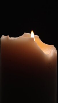 Vertical view of thick cream-colored candle with an irregular, melted rim, burning with a bright flame and surrounded by thick smoke, rotates against a black background.
