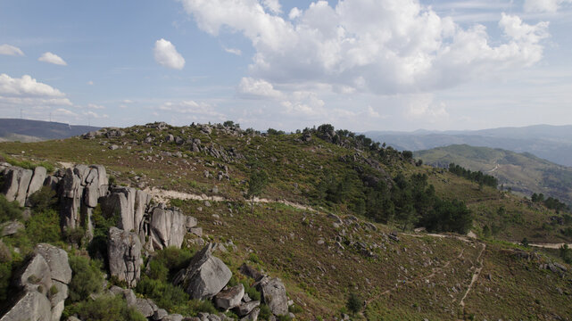 Aerial view of Peneda-Geres National Park featuring rugged granite rock formations, sparse vegetation, pine trees, and a winding dirt path under a blue sky in Portugal.