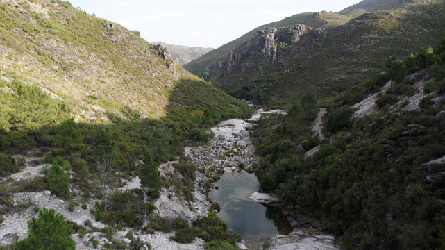 Aerial view of Pocos Verdes Sete Lagoas featuring a clear mountain stream flowing through a rocky valley with green hills and pine trees in Cabril, Vila Real, Portugal.