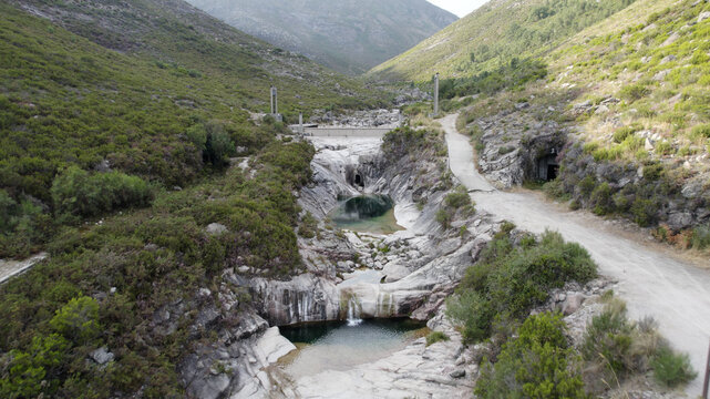 Aerial view of the Pocos Verdes natural pools and waterfalls in a rocky riverbed, surrounded by lush green hills and a dirt path in Cabril, Vila Real, Portugal.
