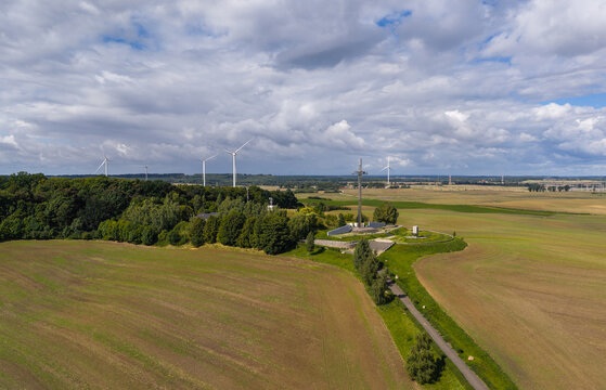 Aerial view of the Pope's Cross monument surrounded by lush trees, agricultural fields, and wind turbines in the distance in Pelplin, Pomeranian Voivodeship, Poland.