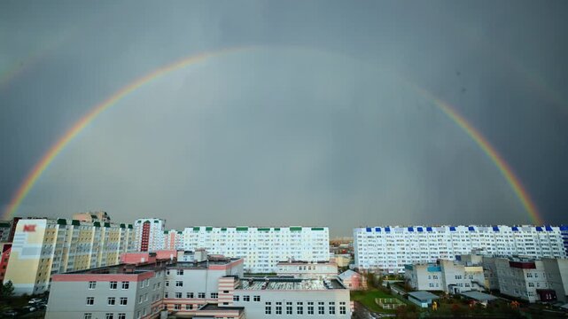 Rainbow arcs over city blocks after stormy skies. Clouds linger above muted, concrete buildings. Bright colors contrast with gray, dreary atmosphere. in this urban landscape