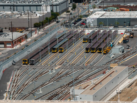 Aerial view shows an urban transit depot in Los Angeles, California, with multiple passenger subway trains parked along parallel tracks beside industrial warehouses and city streets.