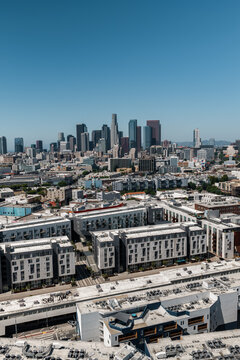 Aerial perspective reveals the urban sprawl and modern residential complexes of the Arts District leading toward the downtown Los Angeles skyscraper skyline under a clear afternoon sky.