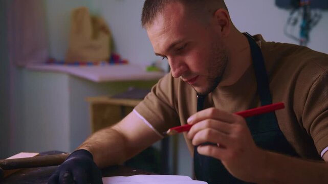 Focused craftsman writes notes with red pen. Dressed in apron, he works intently at wooden table. Soft lighting highlights his concentration. Ideal for DIY, artisan, or workshop projects