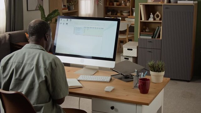 Mature African American man analyzing financial spreadsheets on computer screen and taking notes from rear view. Footage fits remote work campaigns and data analytics software ads